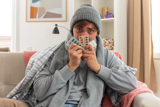 Ängstlicher junger kranker slawischer Mann mit Schal um den Hals mit Wintermütze, der Medizinblisterpackungen auf der Couch im Wohnzimmer hält