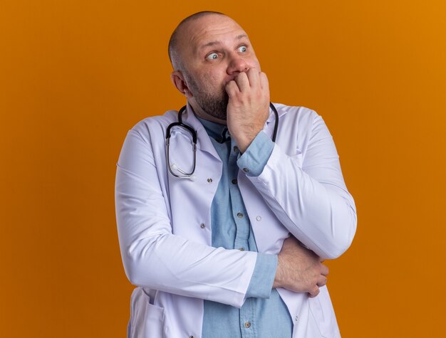 Ängstlicher männlicher Arzt mittleren Alters, der ein medizinisches Gewand und ein Stethoskop trägt und auf die beißenden Finger der Seite schaut, die auf einer orangefarbenen Wand isoliert sind?