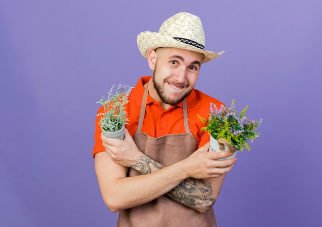 Ängstlicher männlicher Gärtner, der Gartenhut trägt, kreuzt Arme, die Blumentöpfe halten