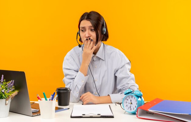 Ängstliches junges Callcenter-Mädchen, das Headset am Schreibtisch sitzt und Seite mit Hand auf Mund lokalisiert auf Orange betrachtet