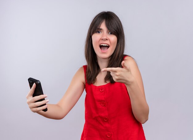 Ängstliches junges kaukasisches Mädchen, das Telefon hält und Finger auf Telefon zeigt Kamera auf lokalisiertem weißem Hintergrund betrachtet