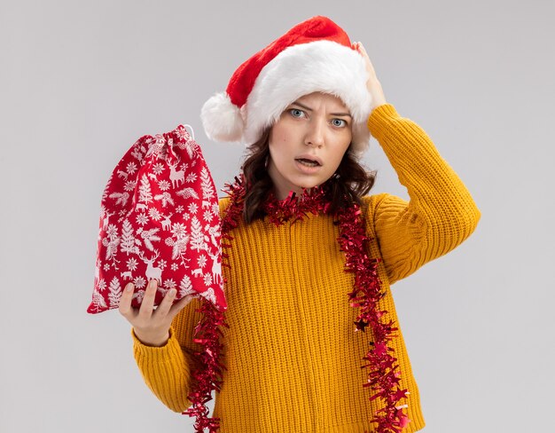 Ängstliches junges slawisches Mädchen mit Weihnachtsmütze und mit Girlande um Hals legt Hand auf Kopf und hält Weihnachtsgeschenkbeutel lokalisiert auf weißem Hintergrund mit Kopienraum