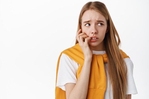 Ängstliches, schüchternes Mädchen, das verängstigt und ängstlich aussieht, in die Finger beißt und nach rechts schaut, sich nervös und verängstigt fühlt und auf weißem Hintergrund steht