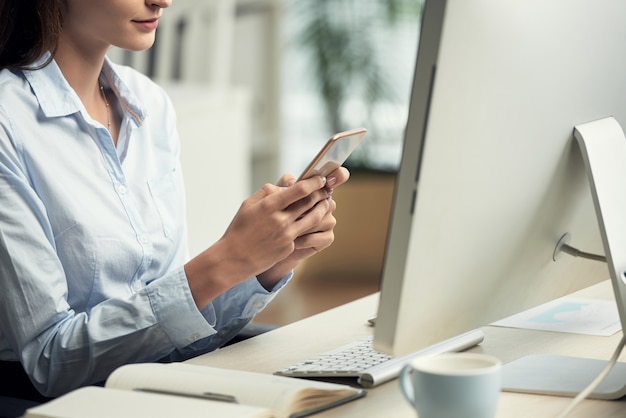 Nicht erkennbare Frau, die im Büro vor Computer sitzt und Smartphone verwendet