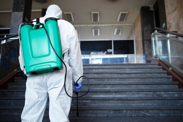 Kostenloses Foto nicht erkennbare person im weißen chemikalienschutzanzug mit reservoir, das den öffentlichen korridor desinfiziert, um die ausbreitung des hoch ansteckenden koronavirus zu stoppen