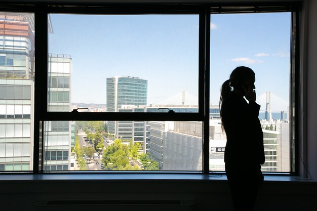 Nicht erkennbare Silhouette der Geschäftsfrau, die nahe Fenster steht und auf Handy spricht. Professioneller Manager in Schatten und Stadtbild. Geschäfts-, Kommunikations- und Unternehmenskonzept