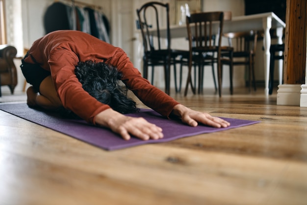 Nicht erkennbarer Mann mit schwarzen Haaren, der zu Hause Yoga macht, sich in Balasana- oder Kinderpose ausruht, die Körpermuskulatur zwischen den Asanas entspannt, den unteren Rücken und die Hüften streckt. Entspannungs- und Gesundheitskonzept