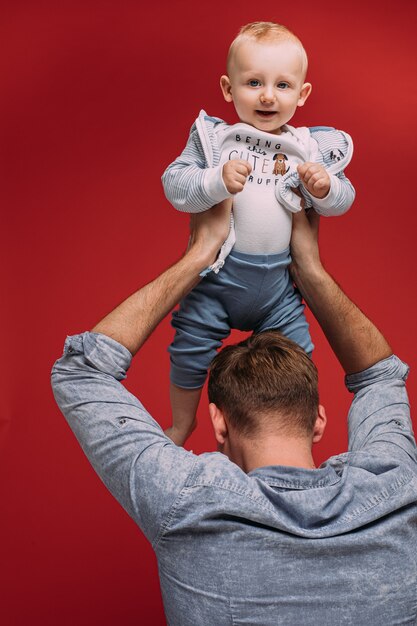 Nicht erkennbarer Vater, der seinen süßen Sohn in den Armen über seinem Kopf vor rotem Hintergrund hält. Lächelndes Baby, das Kamera betrachtet.