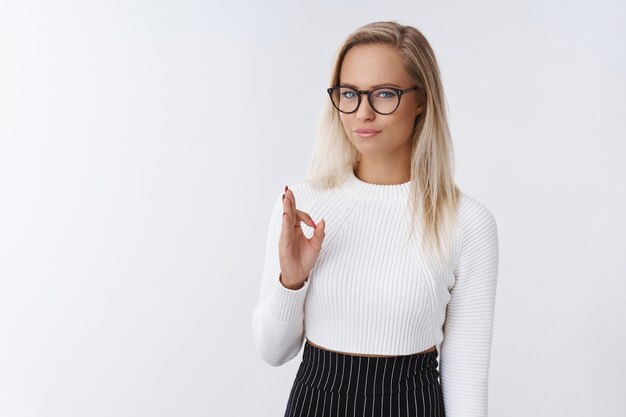 Nicht schlecht genehmigen Ergebnis. Porträt einer begeisterten und zufriedenen, gut aussehenden, stylischen Mitarbeiterin in Brille mit blonden Haaren, die ein Okay-Zeichen zeigt und knifflig lächelt, um eine tolle Auswahl an Freunden zu sehen.