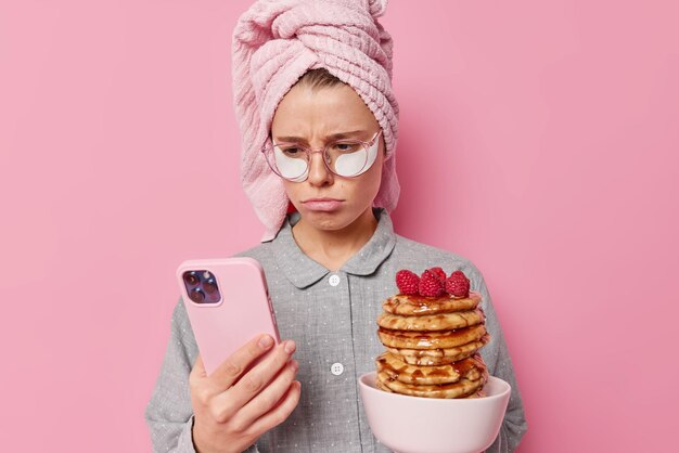 Niedergeschlagene, unzufriedene junge Frau liest schlechte Nachrichten im Internet, scrollt soziale Netzwerke über Smartphone, nachdem das Erwachen eine Schüssel mit Pfannkuchen mit Sirup hält und Schönheitsflecken im Pyjama anwendet