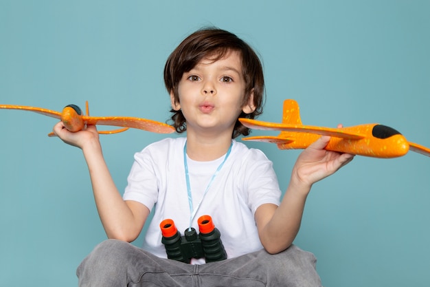niedlicher Junge der Vorderansicht, der orange Spielzeugflugzeuge im weißen T-Shirt auf dem blauen Schreibtisch hält