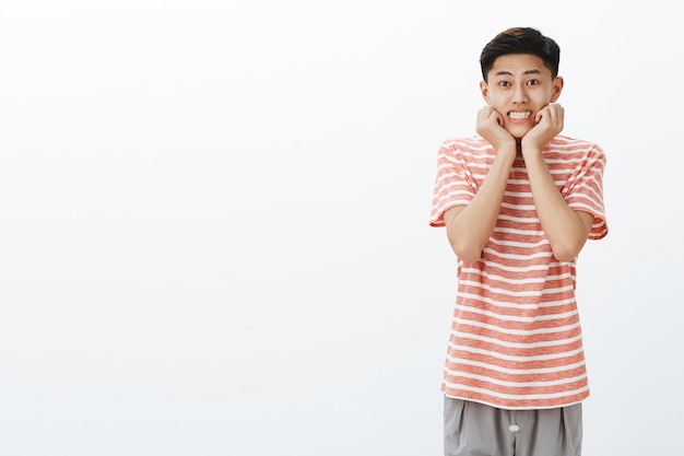 Niedlichkeitsüberlastung. Porträt des dummen attraktiven netten asiatischen Kerls mit dem dunklen kurzen Haar, das breit lehnendes Gesicht auf Handflächen in der glamourösen weiblichen Pose lächelt