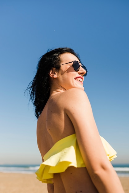 Niedrige Winkelsicht der lächelnden jungen Frau im Bikini, der gegen klaren Himmel am Strand steht