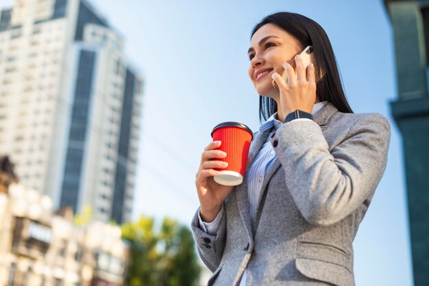 Niedriger Winkel der Smiley-Geschäftsfrau, die am Telefon spricht, während sie Kaffee draußen trinkt