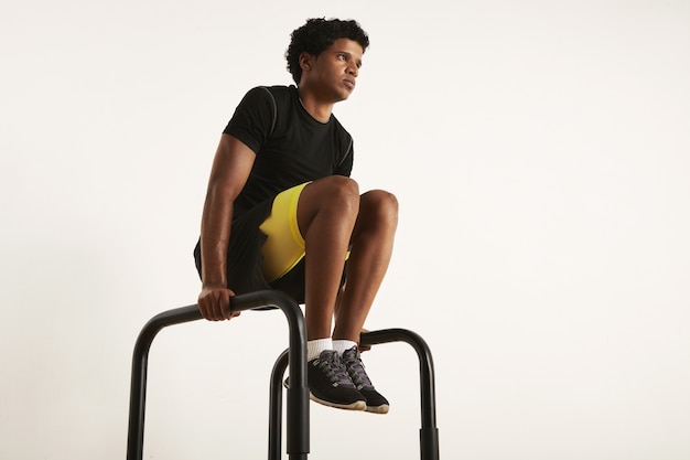 Niedriges Winkelfoto eines starken muskulösen mageren schwarzen männlichen Modells mit einem Afro in der schwarzen Trainingskleidung, die Knie auf den parallelen Balken ansteigend auf Weiß anhebt.