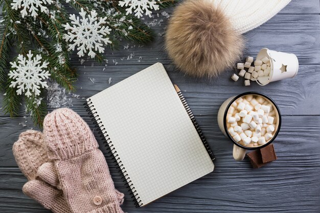 Notebook in der Nähe von Strickhandschuhe, Hut, Tasse mit Marshmallows und Tannenzweig