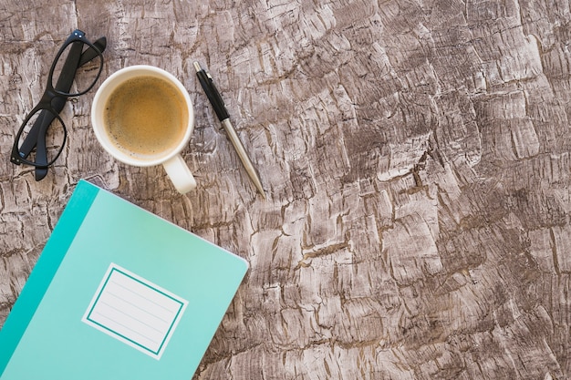 Notizbuch; Kaffeetasse; Brillen und Stift auf strukturierten Hintergrund