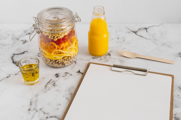 Nudelsalat im Glas mit Öl; Saft; Klemmbrett und Gabel auf Marmoroberfläche