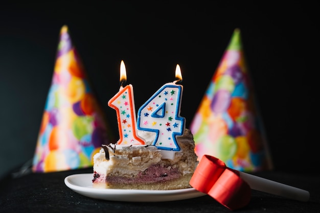 Nummer 14 geburtstag beleuchtete kerze auf dem stück kuchen mit partyhut und partyhorngebläse