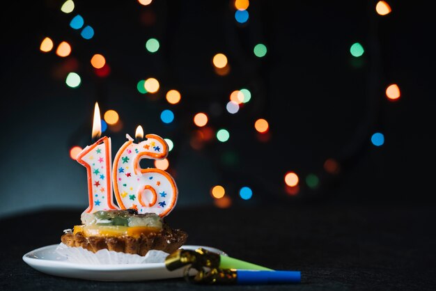 Nummer 16 Geburtstag beleuchtete Kerze auf der Scheibe der Torte mit Partyhorngebläse gegen belichteten Bokeh-Hintergrund