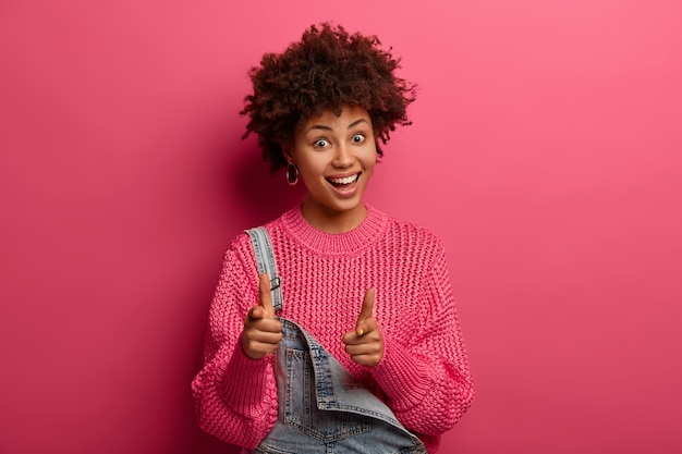 Nur du. Die fröhliche afroamerikanische Frau mit den lockigen Haaren zeigt direkt mit der Geste der Fingerpistole oder der Handfeuerwaffe, hat einen fröhlichen Ausdruck, trägt einen locker gestrickten Pullover, wirbt für Produkt und Verkauf