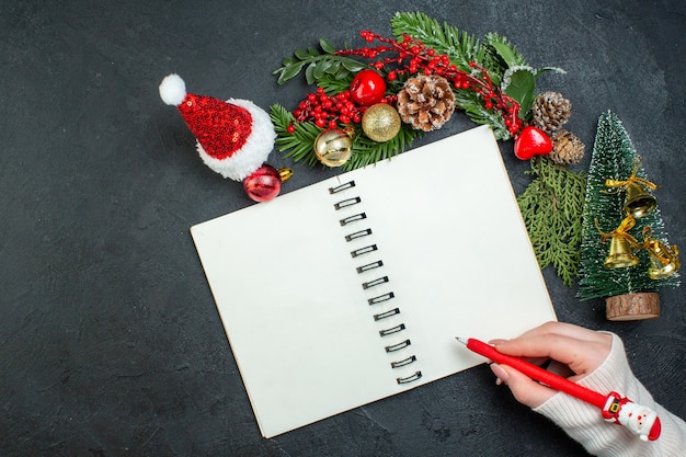 Oben Ansicht der Weihnachtsstimmung mit Tannenzweigen-Weihnachtsbaum-Weihnachtsmannhuthand, die einen Stift auf Spiralblock auf dunklem Hintergrund hält