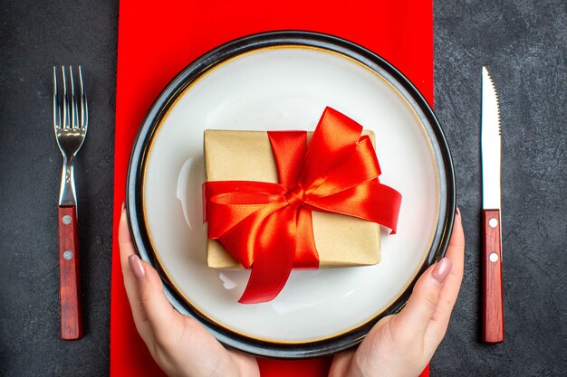 Oben Ansicht des nationalen Weihnachtsmahlzeithintergrundes mit Hand, die leere Teller mit bogenförmigem rotem Band auf einer roten Serviette und Besteck auf schwarzem Tisch hält