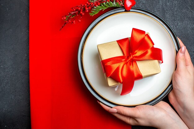 Oben Ansicht des nationalen Weihnachtsmahlzeithintergrundes mit Hand, die leere Teller mit bogenförmigem rotem Band und Tannenzweigen auf einer roten Serviette auf schwarzem Tisch hält