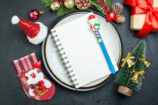 Oben Ansicht des Notizbuchs mit Stift auf Teller Weihnachtsbaum Tannenzweige Nadelbaumkegel Geschenkbox Weihnachtsmann Hut Weihnachtssocke auf schwarzem Hintergrund