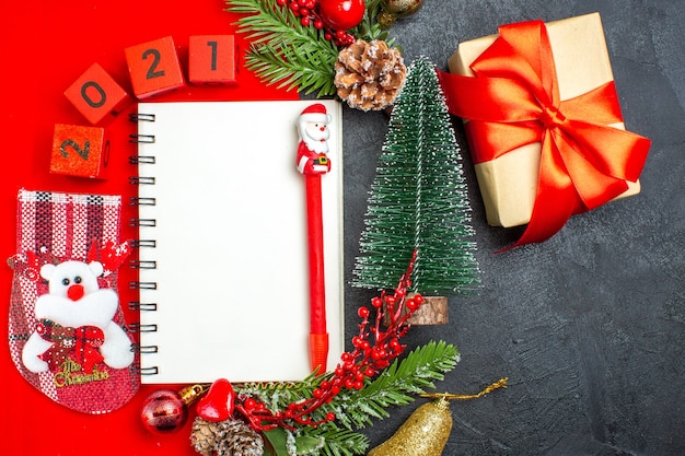 Oben Ansicht des spiralförmigen Notizbuchdekorationszubehörs Tannenzweige-Weihnachtssockenzahlen auf einer roten Serviette und Geschenkweihnachtsbaum auf dunklem Hintergrund