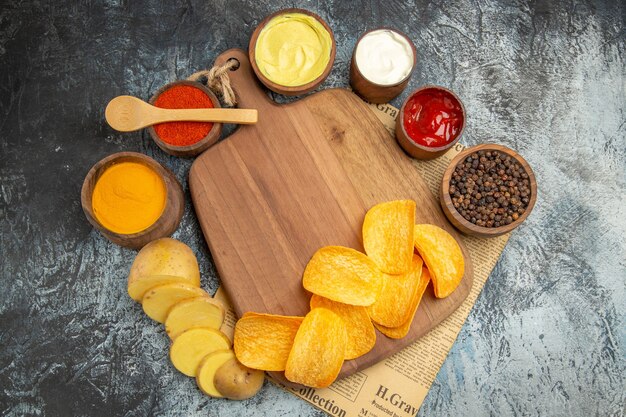 Oben Ansicht von hausgemachten köstlichen Kartoffelchips auf Holzschneidebrett verschiedene Gewürze und Aromen auf Zeitung auf grauem Tisch