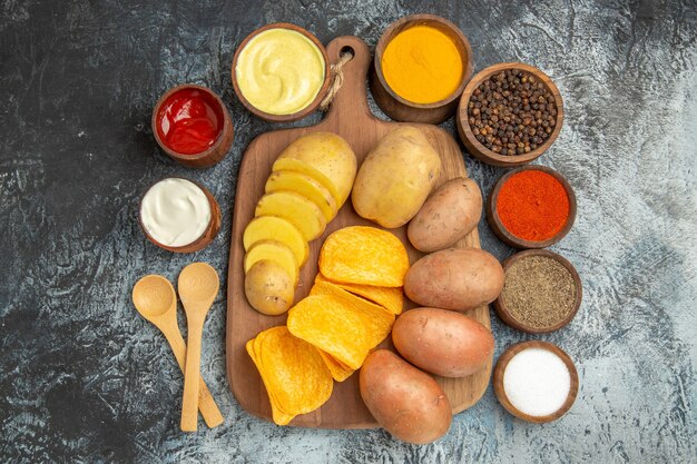Oben Ansicht von knusprigen Pommes und ungekochten Kartoffeln auf Holzschneidebrett und verschiedenen Gewürzen Mayonnaise Ketchup auf grauem Tisch