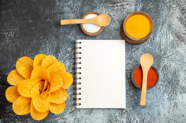 Oben Ansicht von leckeren Kartoffelchips, die wie Blumen gewürzt sind, verschiedene Gewürze mit Löffeln darauf und Notizbuch auf grauem Tisch