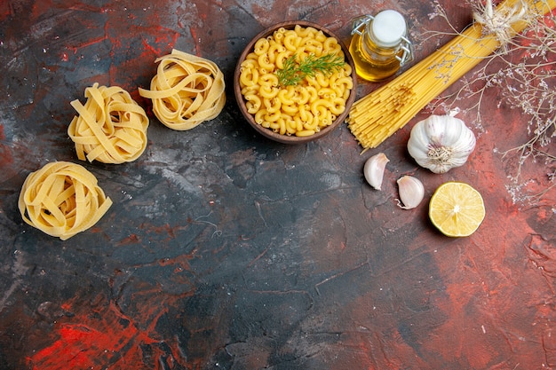 Oben Ansicht von ungekochten drei Portionen Spaghetti und Schmetterlingsnudeln in einer braunen Schüssel und einer grünen Zwiebel-Zitronen-Knoblauch-Ölflasche auf einer gemischten Farbtabelle
