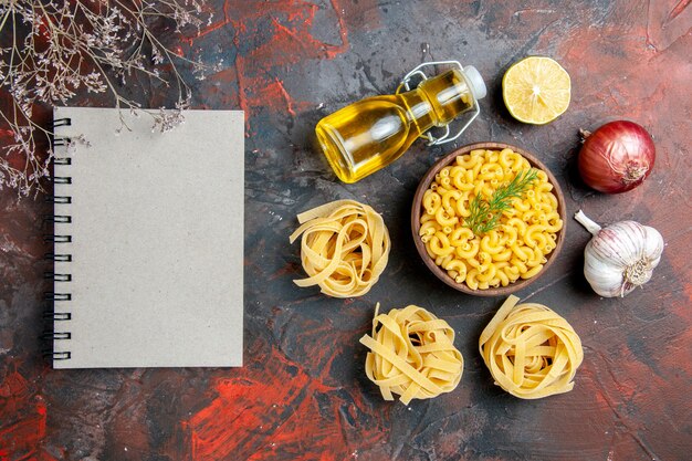 Oben Ansicht von ungekochten drei Portionen Spaghetti und Schmetterlingsnudeln in einer braunen Schüssel und einer Zitronen-Knoblauchöl-Flasche mit Frühlingszwiebeln neben einem Notizbuch auf einer Tabelle mit gemischten Farben