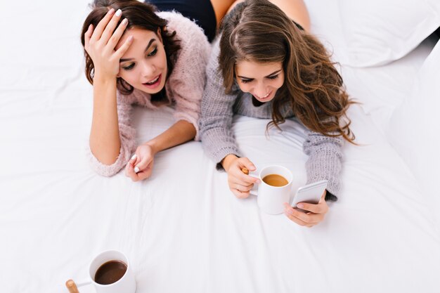 Oben Ansicht zwei junge attraktive Frauen, die Spaß zusammen auf weißem Bett haben. Guten Morgen von hübschen Mädchen, am Telefon im Internet surfen, Tee trinken, lächeln.