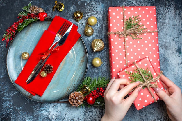 Oben Blick auf die Hand, die eine Geschenkbox und ein Besteck mit rotem Band auf einer dekorativen Serviette auf einem blauen Teller und Weihnachtszubehör und Weihnachtssocke auf dunklem Hintergrund öffnet