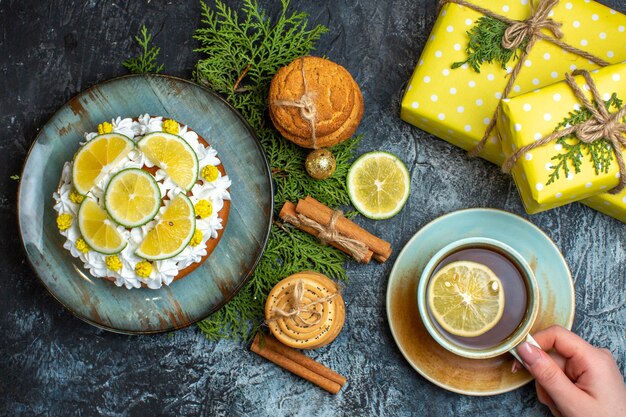 Oben Blick auf die Hand, die eine Tasse schwarzen Tee cremig leckeren Kuchen und Tannenzweige Zitronen-Zimt-Limonen gelbe Geschenkboxen auf dunklem Hintergrund hält