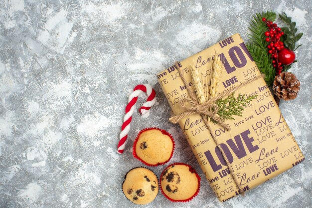 Oben Blick auf ein schönes Weihnachtsgeschenk mit Liebesaufschrift und kleinen Cupcakes Tannenzweigen Dekorationszubehör Nadelbaumkegel auf Eisoberfläche