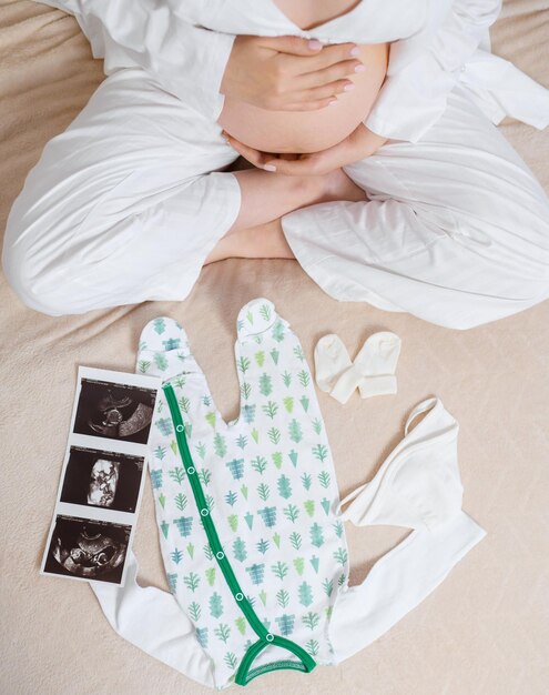 Oben Blick auf eine anonyme Frau in weißem, gemütlichem Pyjama, die auf dem Bett sitzt, mit Ultraschallbild des süßen Bodysuits und der Socken der Gebärmutter, während sie den schwangeren Bauch streichelt und auf ein Baby und einen Teil der Familie wartet