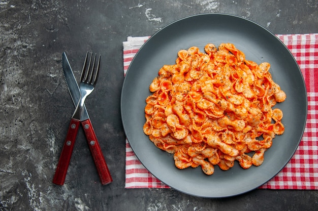 Oben Blick auf einfache Pasta-Mahlzeit zum Abendessen auf einem schwarzen Teller und Besteck auf einem roten, gestreiften Handtuch auf dunklem Hintergrund