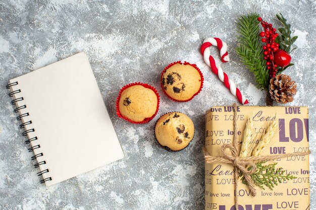 Oben Blick auf geschlossenes Notizbuch und kleine Cupcakes schönes Weihnachtsgeschenk mit Liebesaufschrift und Tannenzweigen Dekorationszubehör Nadelbaumkegel auf der linken Seite auf Eisoberfläche