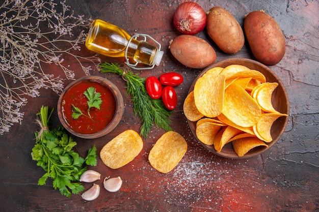 Oben Blick auf hausgemachte köstliche knusprige Kartoffelchips in einer kleinen braunen Schüssel Kartoffeln Ölflaschengrün und Tomaten-Knoblauch-Ketchup auf dunklem Tisch