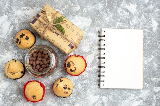 Oben blick auf köstliche kleine cupcakes und schokolade in einem glastopf neben weihnachtsgeschenk und geschlossenem notizbuch auf eisfläche