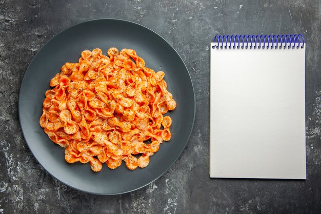 Oben Blick auf köstliche Pasta-Mahlzeit auf einem schwarzen Teller zum Abendessen und Notizbuch auf dunklem Hintergrund