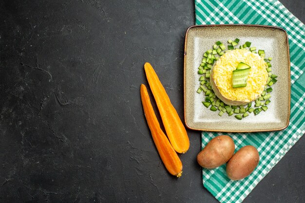 Oben Blick auf köstlichen Salat, serviert mit gehackter Gurke auf halb gefaltetem grün gestreiftem Handtuch und Karottenkartoffeln auf dunklem Hintergrund