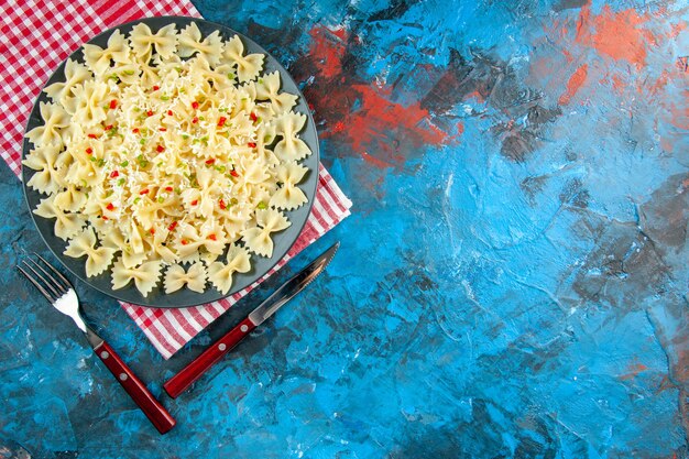 Oben Blick auf rohe italienische Farfalle-Nudeln mit Gemüse auf einem schwarzen Teller auf einem rot gestreiften Handtuch neben dem Besteck auf der rechten Seite auf blauem Hintergrund