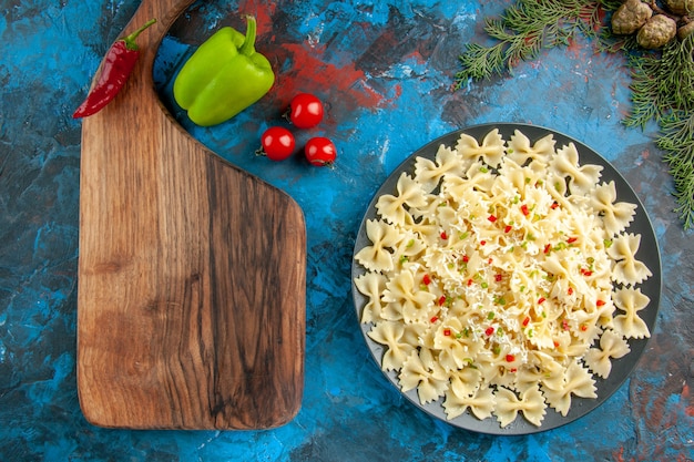 Oben Blick auf rohe italienische Farfalle-Nudeln mit Gemüse auf einem schwarzen Teller und Holzbrett und Gabel neben Paprikatomaten auf blauem Hintergrund