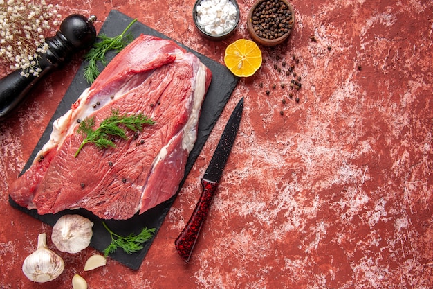 Oben Blick auf rohes frisches rotes Fleisch mit Grün und Pfeffer auf schwarzem Brettmesser Holzhammer Salz Zitrone auf Ölpastellrotem Hintergrund