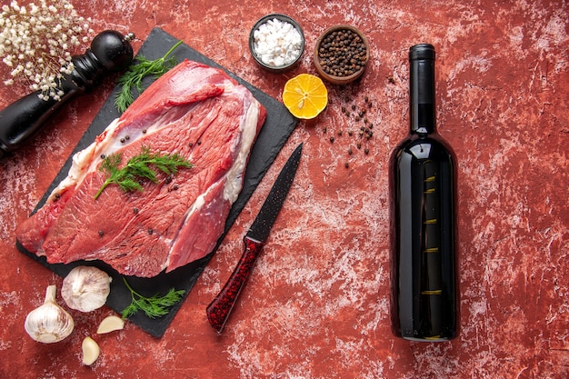 Oben Blick auf rohes frisches rotes Fleisch mit Grün und Pfeffer auf Tafelmesser Knoblauch Zitronengewürze Holzhammer Zitronenweinflasche auf Ölpastellrotem Hintergrund